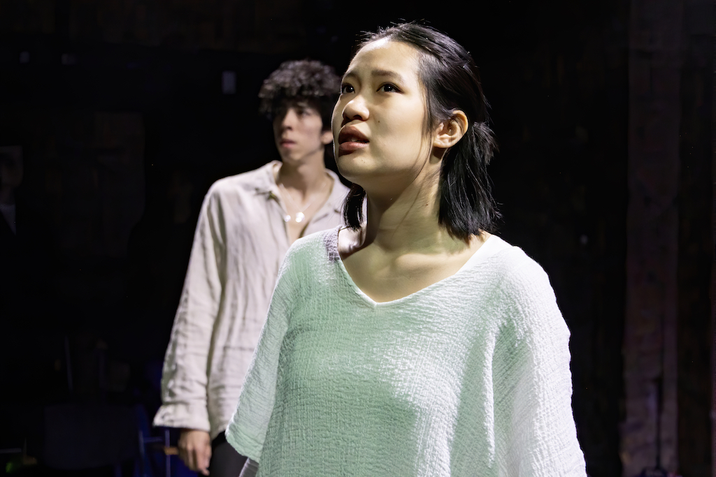 Actor stands and looks up in mid-performance while a second actor stands behind her.